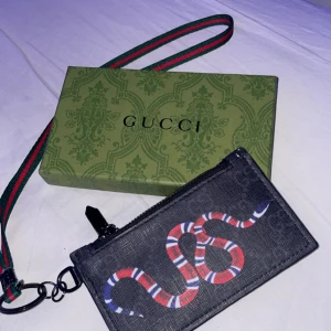 Svart plånbok från Gucci med ormtryck - Snygg svart plånbok från Gucci med ett coolt ormtryck i rött och vitt. Plånboken har flera kortfack och en praktisk dragkedja. Den kommer med en avtagbar rem i klassiska Gucci-färger. Perfekt för att hålla ordning på dina kort och kontanter. Hör av er vid frågor 