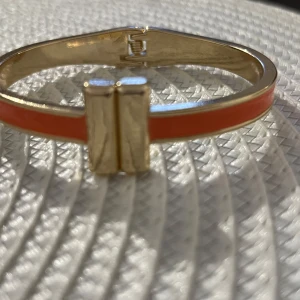 Orange och guld armband - Elegant armband i orange och guld med en stilren design. Armbandet har en slät yta och en dekorativ gulddetalj i mitten som ger en lyxig känsla. Perfekt för att lägga till en färgklick till din outfit.
