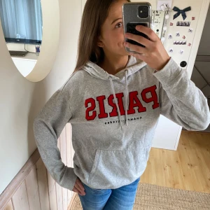 Grå hoodie med Paris-tryck - Säljer en grå hoodie med stort rött 'PARIS' tryck på framsidan. Hoodien har en känguruficka och justerbar dragsko i huvan. Perfekt för en avslappnad stil.