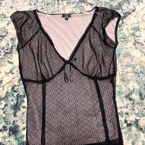 Svart spetsblus från MEXX - Säljer en elegant svart spetsblus från MEXX. Den har korta ärmar och en v-ringad design med knytning framtill. Perfekt för en festlig kväll eller en stilfull dag på stan. Blusen är genomskinlig och ger en chic look.