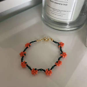 Färgglatt pärlarmband med blommor - Säljer ett superfint armband med små orangea pumpor gjorda av pärlor. Armbandet har en mörkgrön bas och är perfekt för att liva upp vilken outfit som helst. Det har en justerbar kedja i guld för att passa olika storlekar. Perfekt för sommarens alla f