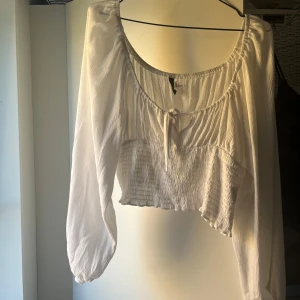 Vit blus från Divided - Säljer en superfin vit blus från Divided. Den har långa ärmar och en söt knytning framtill. Blusen är croppad och har en smockad detalj vid midjan som ger en snygg passform. Perfekt för både vardag och fest!