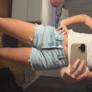 Ljusblå jeansshorts - Säljer ett par ljusblå jeansshorts med råa kanter och klassisk femficksdesign. Shortsen har en knappgylf och är perfekt för sommardagar. De är högmidjade och ger en avslappnad look.