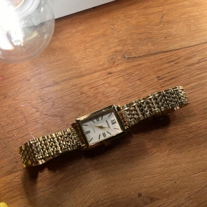 Guldfärgad klocka med rektangulär urtavla - Elegant guldfärgad klocka med rektangulär urtavla och romerska siffror. Armbandet är i metall med en flätad design som ger en lyxig känsla. Perfekt accessoar för att lyfta vilken outfit som helst. 🥂💖