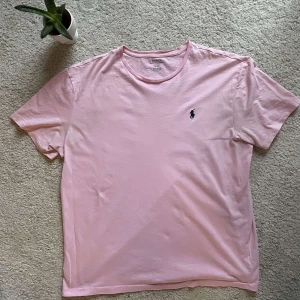 Rosa t-shirt från Polo Ralph Lauren - Säljer en stilren rosa t-shirt från Polo Ralph Lauren med det klassiska broderade logotypen på bröstet. T-shirten är kortärmad och tillverkad i mjuk bomull för en bekväm passform. Perfekt för en avslappnad look.