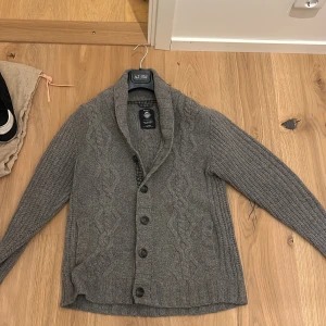 Grå stickad kofta från GANT - Säljer en snygg grå stickad kofta från GANT i 100% lammull. Koftan har ett kabelstickat mönster och knappar framtill. Den har långa ärmar med förstärkta armbågar i ett rutigt mönster. Perfekt för kyliga dagar! Pris kan diskuteras