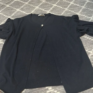 Marinblå kofta från Gina Tricot - Säljer en stilren marinblå kofta från Gina Tricot med en glittrig stjärndetalj framtill. Koftan har långa ärmar med volangdetaljer och är perfekt för en elegant look. Passar bra till både vardag och speciella tillfällen.