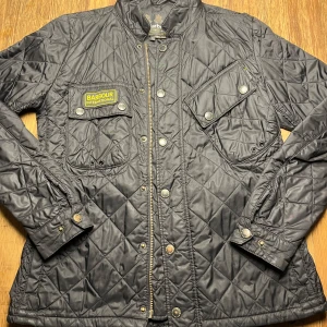 Svart quiltad jacka från Barbour - Säljer en snygg svart quiltad jacka från Barbour International. Jackan har en stilren design med knappar och dragkedja framtill samt två bröstfickor med knappar. Perfekt för en cool och avslappnad stil.