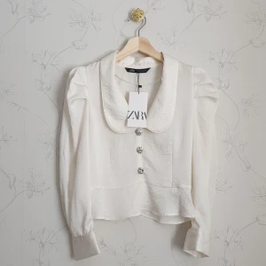 Zara vit blus med strass & puffärmar  - 🧁FÖRST TILL KVARN!🧁En vit slutsåld blus från Zara, helt ny & aldrig använd. Blusen är en aning skir med rundad krage. Knapparna är även av strass och blusen har även fina puffärmar. Det var ett impuls köp, nypris 359