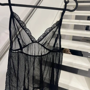 Svart spetslinne - Säljer ett elegant svart spetslinne med tunna axelband och vacker spetsdetalj vid halsringningen. Linnet är transparent och har en luftig passform, perfekt för lager-på-lager-stil.
