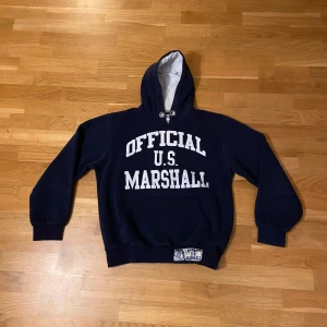 Mörkblå hoodie från U.S. Marshall - Säljer en mörkblå hoodie från U.S. Marshall med vit text på framsidan. Den har en klassisk känguruficka och justerbar huva. Riktigt snygg passform, passar small.