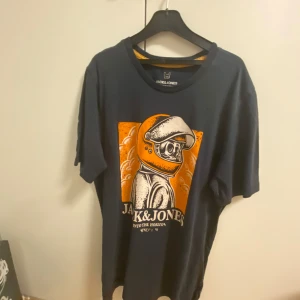 Svart t-shirt från Jack & Jones - Cool svart t-shirt från Jack & Jones med ett unikt tryck av en astronaut i orange och vitt. Perfekt för en avslappnad stil med en touch av rymdäventyr. T-shirten har korta ärmar och en rund halsringning.