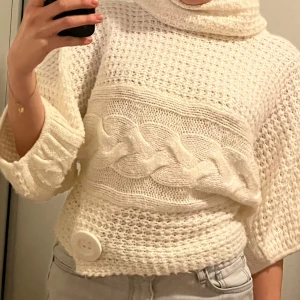 Vit stickad polotröja  - Säljer en mysig vit stickad polotröja med en stor dekorativ knapp framtill. Tröjan har ett flätmönster över bröstet, breda ballongärmar och en hög krage. Storlek S/36. 