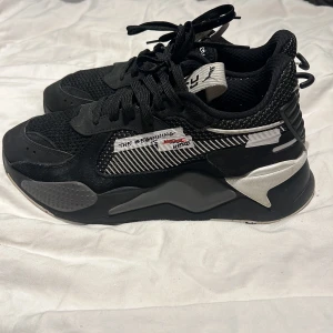 Svarta Puma sneakers med vita detaljer - Snygga svarta sneakers med vita och silverfärgade detaljer. Skorna har snörning och en modern design med textdetaljer på sidan. Perfekta för en stilren look.