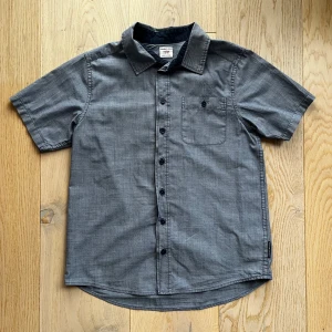 Grå kortärmad skjorta från Polarn O. Pyret - Snygg grå kortärmad skjorta från Polarn O. Pyret i 100% bomull. Skjortan har en klassisk krage och knappar framtill samt en bröstficka med knapp. Perfekt för en stilren och bekväm look.