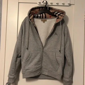 Burberry hoddie  - Säljer en stilren grå huvtröja från Burberry med dragkedja och broderad logga. Insidan av huvan har det klassiska Burberry-rutiga mönstret. Perfekt för en casual look.