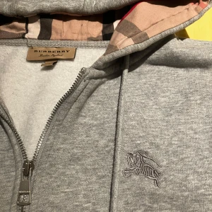 Grå hoodie från Burberry - Säljer en stilren grå hoodie från Burberry med dragkedja och klassiskt rutigt mönster i huvan. Tröjan har långa ärmar och en broderad logga på bröstet. Perfekt för en avslappnad och trendig look. Förpackningen finns kvar.                  Bara att lägga bud!