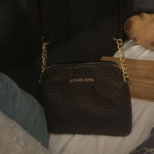 Brun axelväska från Michael Kors - Snygg brun axelväska från Michael Kors med ett diskret mönster och guldfärgade kedjedetaljer. Väskan har en dragkedja upptill och är perfekt för att bära dina nödvändigheter med stil.