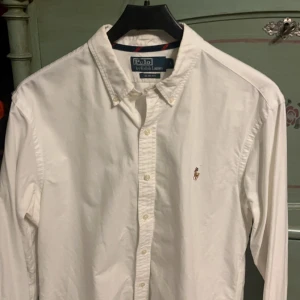 Vit skjorta från Polo Ralph Lauren - Snygg vit skjorta från Polo Ralph Lauren i slim fit. Skjortan har långa ärmar och klassiska knappar framtill. Den är broderad med den ikoniska Polo-loggan på bröstet. Perfekt för en stilren look.