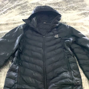 Haglöfs jacka - Säljer en svart dunjacka från Haglöfs. Jackan har en dragkedja framtill och är perfekt för kyligare väder. Den är designad med långa ärmar och en hög krage för extra värme. Passar bra för både vardag och utomhusaktiviteter.