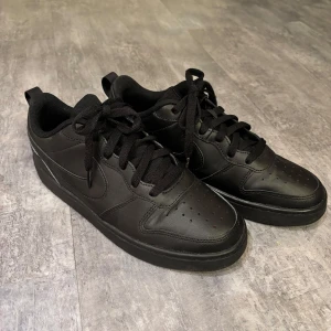 Svarta sneakers från Nike - Säljer ett par helsvarta Nike sneakers med klassisk design och snörning. Skorna har en stilren look med en diskret Nike-logga på sidan. Perfekta för en casual stil.