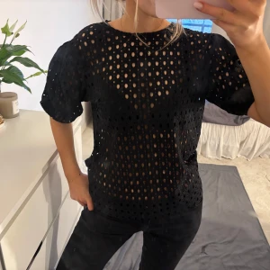 Svart blus från Lindex - Säljer en svart blus från Lindex med ett genombrutet mönster. Blusen har korta puffärmar och en rund halsringning. Perfekt för att lägga till en touch av stil till din garderob.