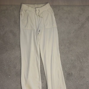 Beige mjukisbyxor från Juicy Couture - Snygga beige mjukisbyxor från Juicy Couture med dragsko i midjan och fickor med knappar bak. Perfekta för en avslappnad stil med en touch av glamour.