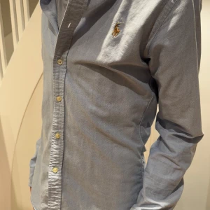 Blå skjorta från Ralph Lauren - Snygg blå skjorta från Ralph Lauren i slim fit. Skjortan har långa ärmar och knappar framtill. Perfekt för en stilren look.