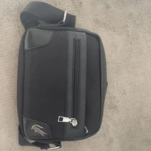 Svart axelväska från Lacoste - Snygg svart axelväska från Lacoste med dragkedja framtill. Väskan har en justerbar axelrem och det ikoniska Lacoste-märket i hörnet. Perfekt för att bära dina dagliga nödvändigheter med stil.🐊🐊