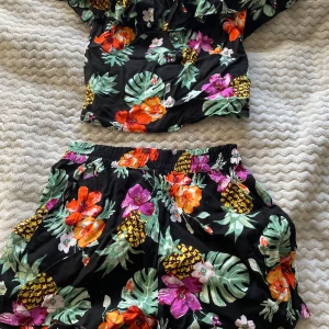 Tropisk offshoulder topp från H&M - Säljer en färgglad offshoulder topp från H&M med tropiskt blommönster i orange, lila och grönt på svart bakgrund 🌺. Den har volangdetaljer och är perfekt för sommardagar. Matcha med shorts för en komplett look! Skriv gärna innan köp😊