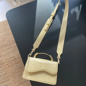 Beige axelväska med krokodilmönster - Säljer en superfin ljusgul axelväska från Hvisk. knappt använd och i jättefint skick. Den har ett justerbart axelband och ett litet handtag för olika bärstilar. Perfekt för både vardag och fest!