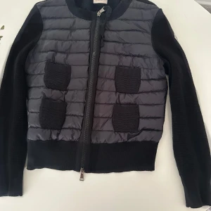 Svart Cardigan jacka Moncler M - Säljer en snygg svart cardigan från Moncler. Den har en quiltad design med stickade detaljer och dragkedja framtill. Perfekt för höst och vinter! Jackan har långa ärmar och två praktiska fickor på framsidan, har en defekt 