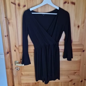 Svart omlottklänning - Säljer en elegant svart omlottjumpsuit med långa ärmar som ser ut som en klänning. Jumpsuiten har en v-ringad design och är perfekt för både vardag och fest. Den är gjord i ett mjukt och bekvämt material som ger en smickrande passform.