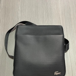 Lacoste axelväska - Lacoste axelväska som är väldigt smidig om du har mycket grejer i fickorna. Totalt 4 olika utrymmen att lägga sina saker i, skick 10/10 knappt använd! Hör av dig vid intresse🙌