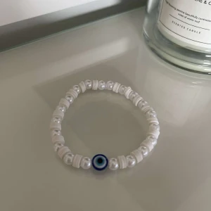 Vitt pärlarmband med öga - Säljer ett snyggt vitt pärlarmband med ett coolt blått öga i mitten. Armbandet är elastiskt och passar perfekt för både vardag och fest. Det är enkelt men ändå unikt med sina vita pärlor och det iögonfallande ögat.