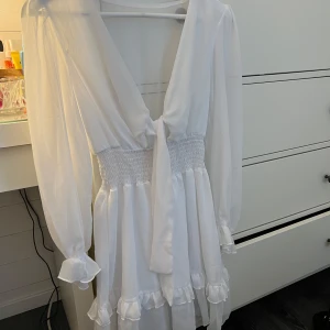 Vit klänning med volanger - Säljer en superfin vit klänning med långa ärmar och volanger. Den har en v-ringad design med knytning framtill och en smockad midja som ger en snygg passform. Perfekt för skolavslutning och student. Den är enbart testad, aldrig använd.