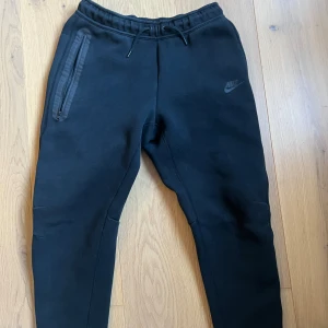 Svarta mjukisbyxor från Nike - Säljer ett par svarta mjukisbyxor från Nike. De är superbekväma och perfekta för chill dagar eller träning. Byxorna har en elastisk midja med snörning och praktiska dragkedjefickor på sidorna. Nike-loggan är diskret placerad på ena benet. Perfekta för både vardag och träning!