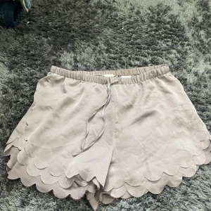 Beige shorts med volangdetaljer - Säljer ett par supersöta beige shorts från H&M i storlek L. De har en elastisk midja med knytband och fina volangdetaljer längs kanterna. Perfekta för sommardagar eller en avslappnad vårlook. Materialet är mjukt och bekvämt, vilket gör dem till ett måste i garderoben!