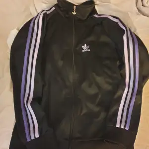 Säljer en snygg svart Adidas jacka med lila ränder längs ärmarna. Jackan har dragkedja och det klassiska Adidas-loggan på bröstet. Perfekt för en sportig look eller bara för att chilla i. Passar både till vardags och träning! 