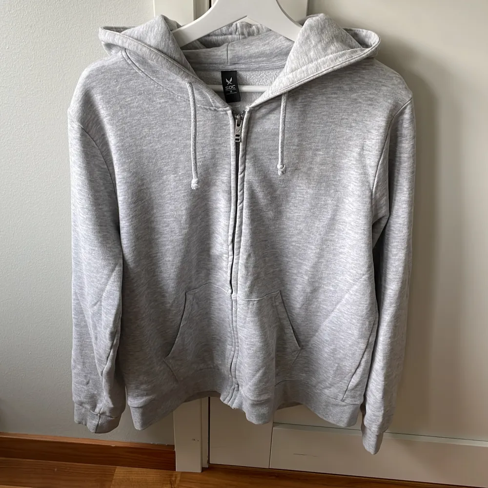 Säljer en grå hoodie från SOC i storlek M. Den är supermjuk och perfekt för både chill och träning. Hoodien har en dragkedja framtill och en skön huva med dragsko. Den har också praktiska fickor för att hålla händerna varma. Perfekt för höst och vår!. Tröjor & Koftor.