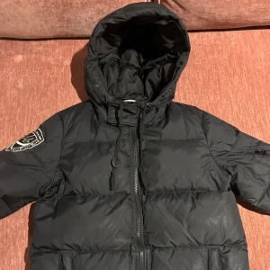 Svart pufferjacka med huva - Säljer en svart pufferjacka med huva från db då den aldrig kommer till användning längre. Jackan har en dragkedja framtill, en innerficka och praktiska fickor med dragkedjor. Perfekt för kalla dagar!