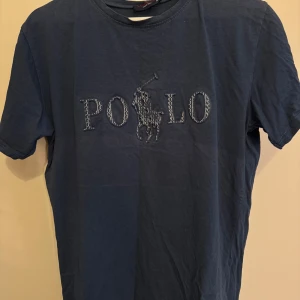 Mörkblå t-shirt från U.S. Polo Assn. - Säljer en snygg mörkblå t-shirt från U.S. Polo Assn. med ett coolt broderat POLO-märke på framsidan. T-shirten är i bomull och har en normal passform. Perfekt för en avslappnad stil!