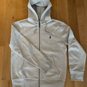Polo Ralph Lauren tech zip - Storlek small. Hoodien har lite noppor på fickorna fram. Dock omöjligt att undvika då den typ är 50/50 bomull och polyester. De ska även sitta metallpluppar på ändarna av snörena som har lossnat. Utöver det, inga fläckar eller andra defekter.