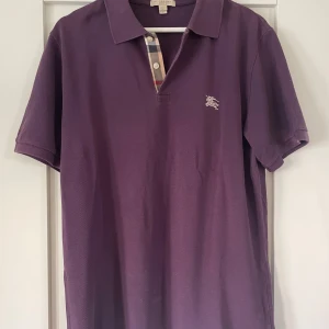 Burberry piké - Burberry piké i storlek M som är lila. Skick 8/10 då den har en liten läck på ärmen och magen.