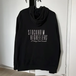 Svart hoodie med tryck - Säljer en svart hoodie med text på ryggen. Den har en dragkedja framtill och en praktisk huva. Oklart vilket märke men bra skick och stilren!