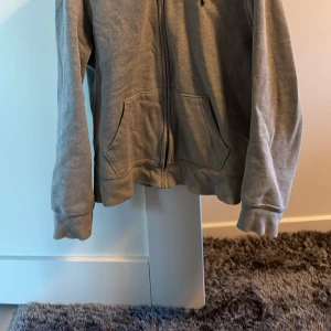 Grå hoodie från Polo Ralph Lauren - Säljer en stilren grå hoodie från Polo Ralph Lauren med dragkedja och broderad logga på bröstet. Tröjan har långa ärmar och en praktisk huva. Perfekt för en avslappnad look. Barn storlek L: 14-16 (dock liten i storlek, passar någon ca 150cm). Bra skick! Pris går att diskutera. 