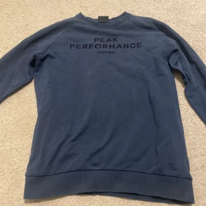 Blå Peak Performance - Säljer en stilren blå sweatshirt från Peak Performance med text på bröstet. Tröjan har långa ärmar och en rund halsringning. Perfekt för en avslappnad stil.