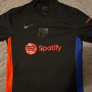 FC Barcelona 24/25  - Säljer den senaste FC Barcelona 24/25 Away Match-tröjan från Nike i svart. En stilren och modern design med de ikoniska blå och röda detaljerna som representerar klubben.  ✔ Storlek: M ✔ Färg: Svart med blå/röda detaljer ✔ Skick: Helt ny ✔ Material: Lätt och ventilerande Dri-FIT-teknologi som håller dig sval och bekväm ✔ Nypris: Standardversionen kostar ca 1 249 kr, medan den autentiska matchversionen är dyrare  Skicka ett meddelande vid intresse. Snabb affär = snabb leverans!