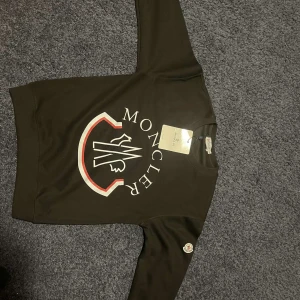 Svart sweatshirt från Moncler - Säljer en stilren svart sweatshirt från Moncler med stort tryck på framsidan. Tröjan har rund hals och långa ärmar med Moncler-logga på ärmen. Perfekt för en casual look.