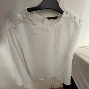 Vit blus med knappar på axlarna - Elegant vit blus från Zara Basic med dekorativa knappar på axlarna. Blusen har en rund hals och långa ärmar, vilket ger en stilren look. Perfekt för en chic och minimalistisk stil.